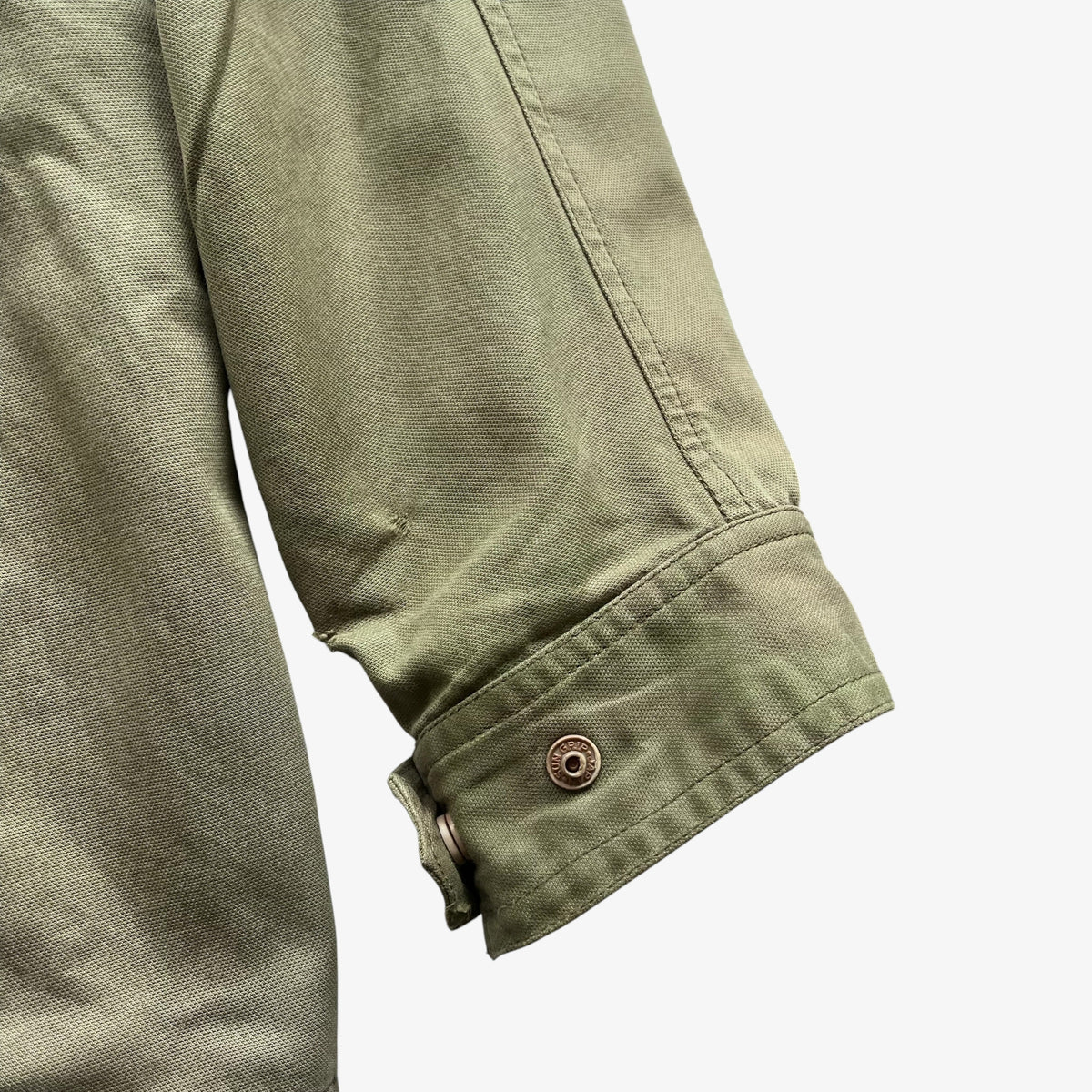 Close-up detail of an olive green fabric garment sleeve with a buttoned cuff, shown against a plain light grey background.