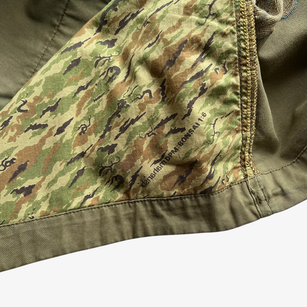 Close-up detail of a Maharishi military jacket's interior lining featuring a green, brown, and black camouflage pattern, finished with a gold-toned zipper and olive green fabric trim.