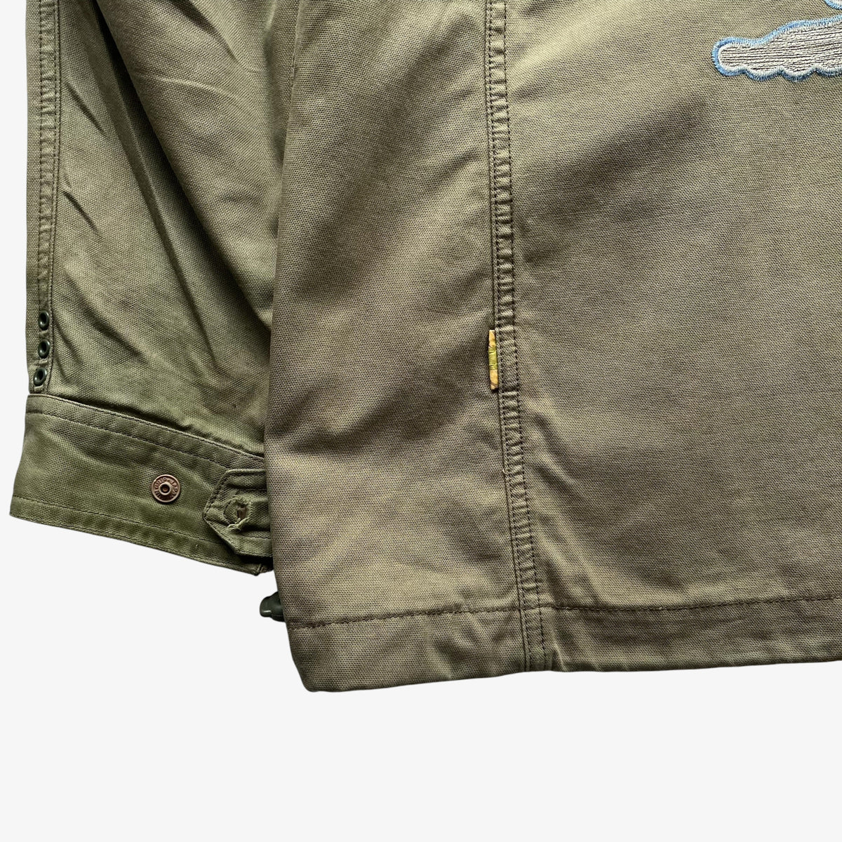 Close-up of an olive green jacket or overshirt showing the sleeve cuff with snap buttons, a vertical seam, a small yellow tab, and part of a light blue embroidered logo on the chest.