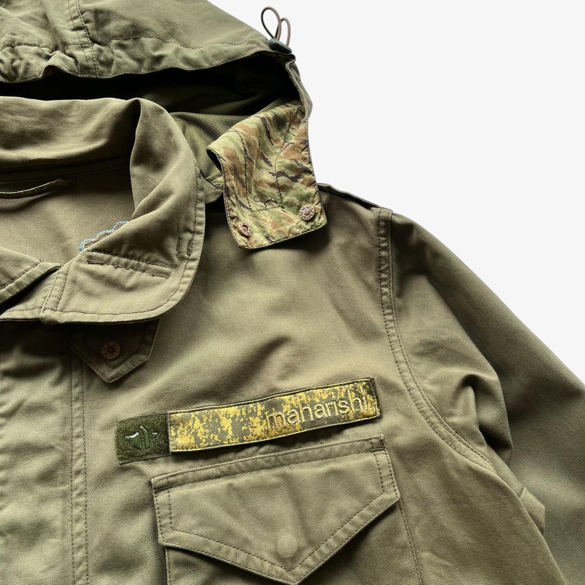 Close-up of a khaki green military-style jacket with a camouflage-patterned collar, a chest pocket, and a visible Maharishi brand patch on the pocket flap.