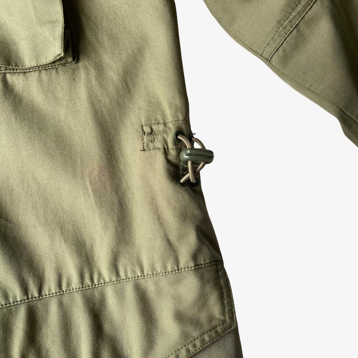 Close-up of an olive green utility jacket showing a side pocket with a metal toggle closure and clean stitching on a white background.