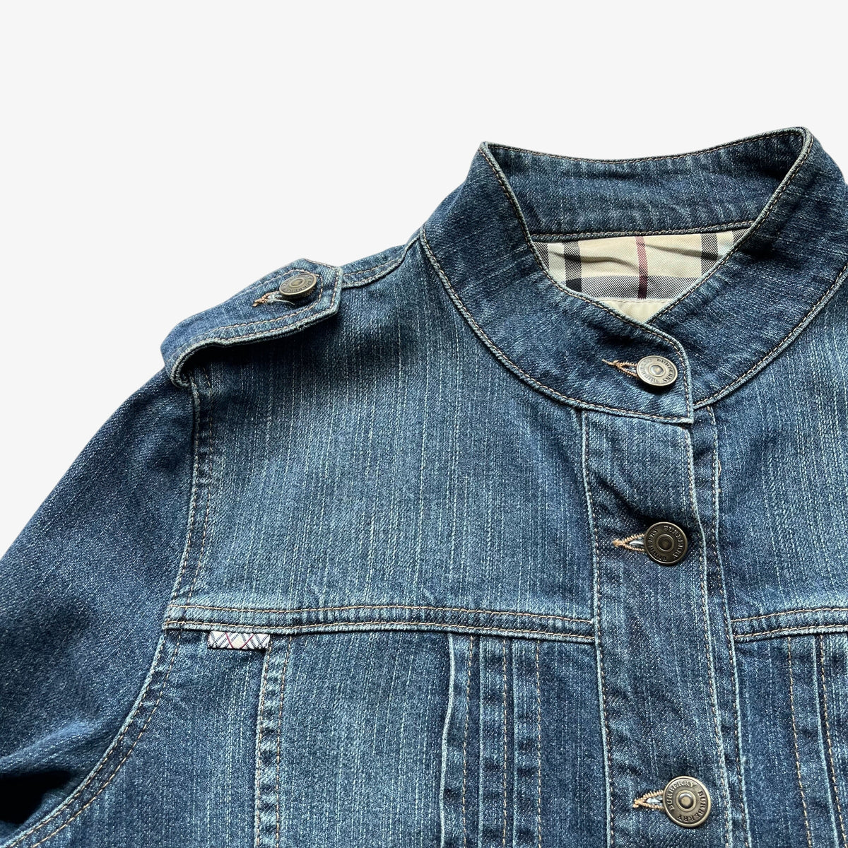 Close-up of a women's Burberry blue military denim jacket with a high stand collar, dark metal snap buttons, and a plaid lining visible inside the collar, set against a plain white background.