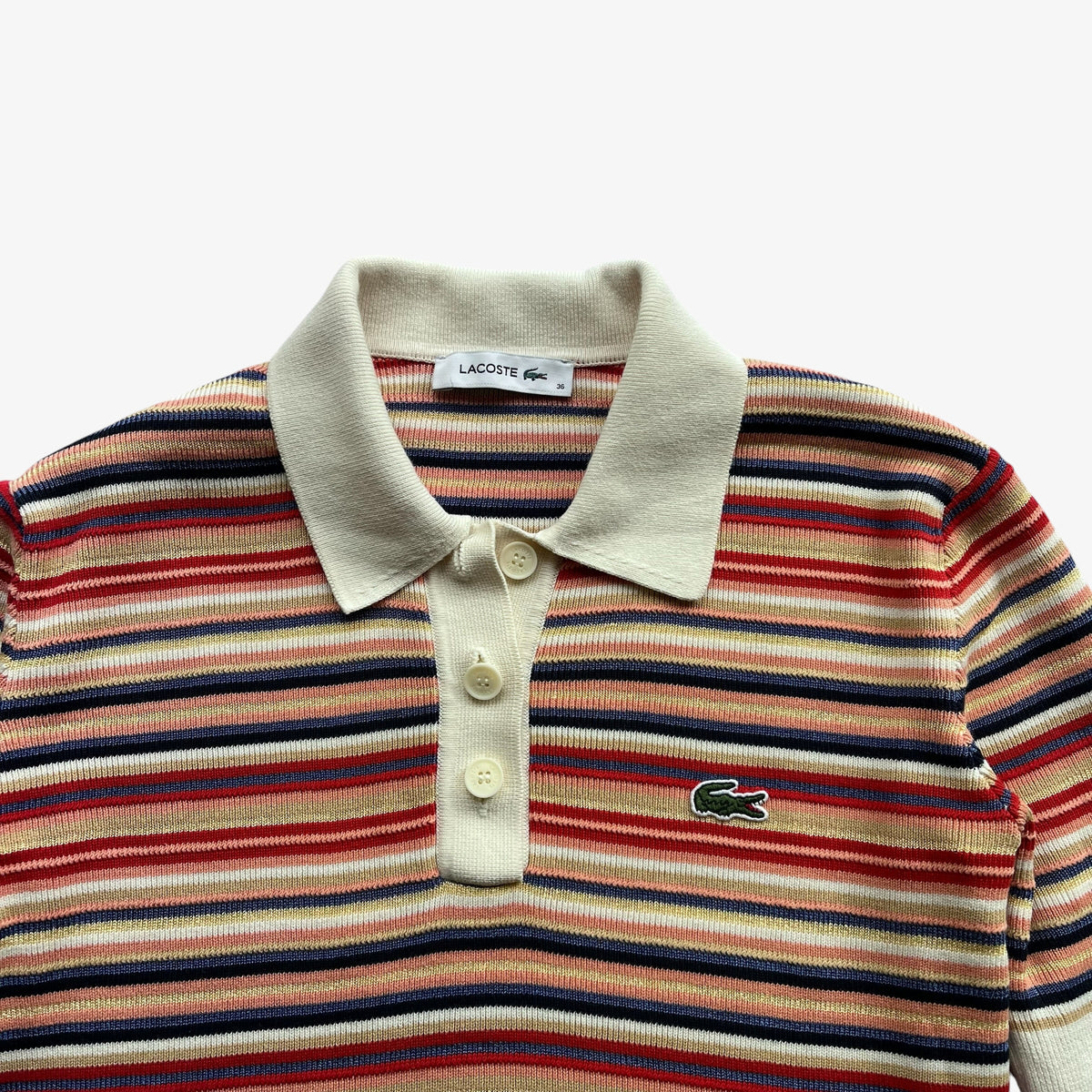 A close-up of a Lacoste striped knit polo shirt featuring a cream collar and placket, with horizontal stripes in red, navy, and beige, and the signature green crocodile logo on the chest.