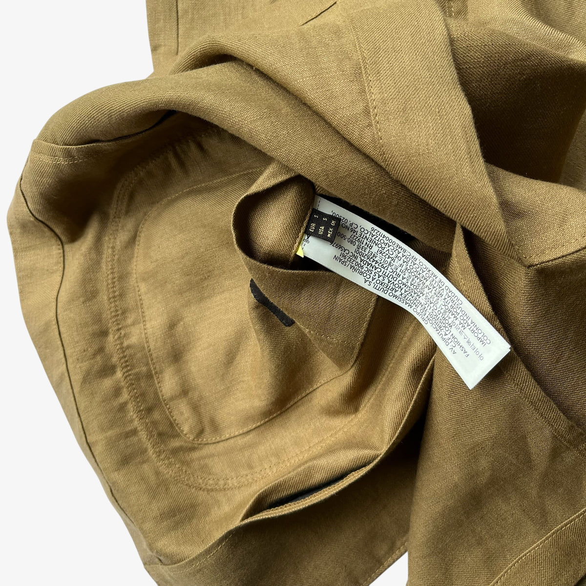 Close-up of a brown women's blazer with a stitched pocket, seam, and a white interior care tag against a plain light background.