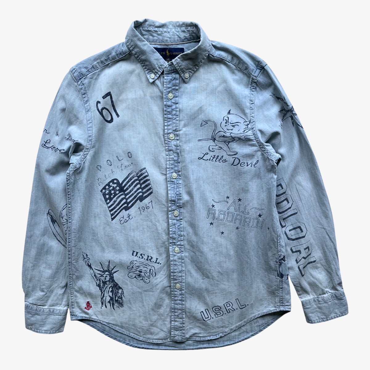 A light blue denim women's Ralph Lauren button-up shirt with various graphic prints including an American flag, the Statue of Liberty, 'Little Devil', and '67' on a white background.
