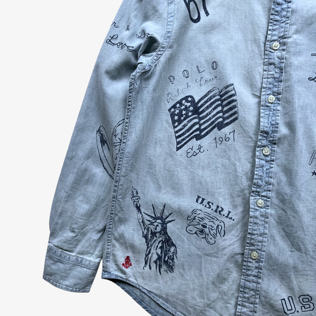 A close-up of a light blue denim button-up shirt featuring black graphic prints of the American flag, Statue of Liberty, and 'U.S.